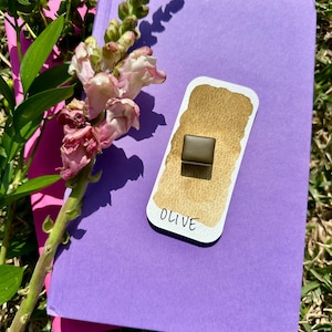 May include: A watercolor swatch labeled "OLIVE" with a small, square, olive-colored pigment. The swatch is on a purple notebook with a pink page peeking out. A sprig of pink and white flowers and green leaves are in the background.