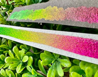 May include: Two swatches of iridescent glitter paint on a gray and white surface. The swatches are a gradient of green, yellow, and pink.