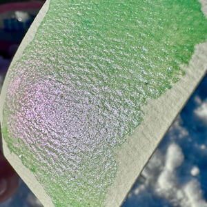 May include: A close-up of a green and iridescent shimmery paint swatch on a white textured surface.