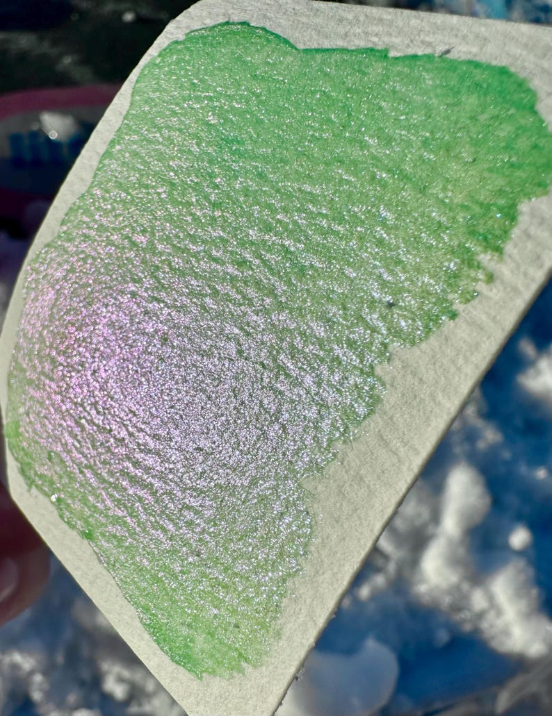 May include: A close-up of a green and iridescent shimmery paint swatch on a white textured surface.