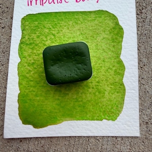 May include: A square, dark green watercolor paint block sits on a white paper with a bright green watercolor wash. The words "impulse buy" are written in pink above the green wash.