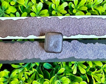 Puede incluir: Una cuenta de vidrio cuadrada, gris oscuro, se encuentra entre dos tiras de papel texturizado, brillante, morado oscuro con bordes rasgados. El fondo es una densa disposición de pequeñas hojas de color verde brillante.