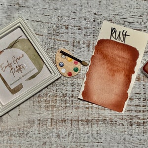K&ouml;nnte beinhalten: Eine Aquarell-Farbpalette mit dem Wort "Rust" darauf. Die Farbe ist rotbraun. Die Palette liegt auf einer Holzoberfl&auml;che. Die Farbe stammt von Emily Grace Palettes, einem Unternehmen f&uuml;r handgemachte Aquarellfarben.