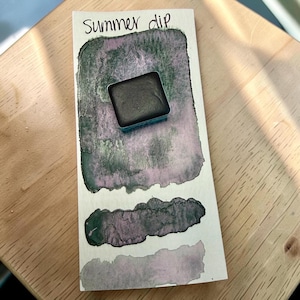 May include: A rectangular watercolour paint pan in a dark metallic green hue resting on a white paper swatch card. The card features the handwritten text Summer dip and displays three distinct painted samples showing colour gradients of green and purple.