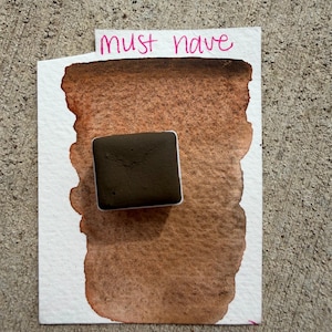 May include: A watercolor swatch on white paper with the words "must have" in pink. The swatch is a blend of brown hues, with a dark brown square watercolor pan on top.