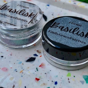 May include: Two small jars of iridescent glitter, one labeled "2 Grams Basilisk Emily Grace Palettes" and the other labeled "1 Gram Basilisk Emily Grace Palettes".