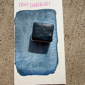 May include: A watercolor paint swatch on white paper with the text "cart confession" in pink. The paint is a dark blue-grey color, with a square of the paint on top of the swatch. The background is a textured, light brown surface.