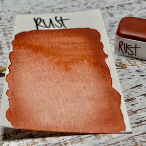 K&ouml;nnte beinhalten: Ein kleiner, quadratischer Beh&auml;lter mit Aquarellfarbe mit der Aufschrift "Rost". Die Farbe ist ein tiefes, r&ouml;tlich-braunes Braun.