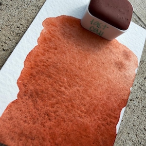 May include: A watercolor paint swatch in shades of orange and brown on textured white paper. A square paint pan with the text "last call" in green is positioned above the swatch, indicating the paint color.