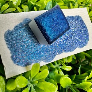 May include: A small square container of blue glitter paint with a white label. The paint is a deep blue with a shimmery finish. The container is sitting on a white surface with a small amount of the paint spilled out.