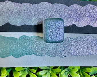 Puede incluir: Un pequeño recipiente cuadrado de pintura de purpurina de color turquesa y morado iridiscente. La pintura está sobre una superficie blanca con una superficie negra detrás.