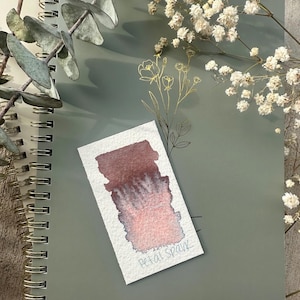 May include: A spiral-bound notebook with a sage green cover, featuring a watercolor swatch labeled "Petal Spark" in shades of pink and burgundy. The image is adorned with sprigs of dried flowers and eucalyptus.