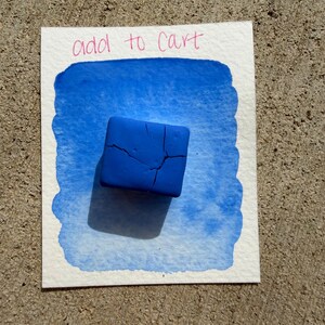 Op de afbeelding: Een vierkant, kobaltblauw object met scheuren ligt op een aquarel. De schildering heeft een blauwe aquarel wassing met de woorden "add to cart" in roze. De achtergrond is een getextureerd, grijs oppervlak.