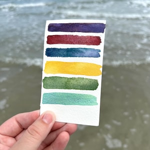 May include: A small white card with seven horizontal watercolor paint swatches in shades of purple, red, blue, yellow, green, and teal. The card is held in front of a blurred ocean background.