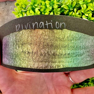 May include: A black card with the word "divination" written in white on the top. The card has a shimmery, iridescent surface with a rainbow of colors.