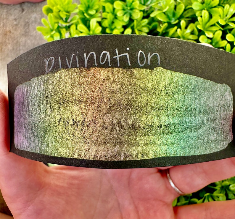May include: A black card with the word "divination" written in white on the top. The card has a shimmery, iridescent surface with a rainbow of colors.