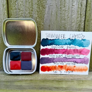 Op de afbeelding: Een open zilveren metalen doos met vier vierkante aquarelverfblokken in tinten donkerblauw, bordeauxrood, rood en paars. Een kaart toont aquarelkleurstalen met de tekst "Summer Chaos" en kleurnamen.