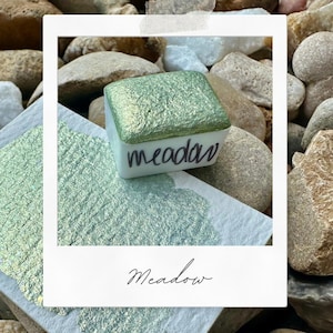May include: A small square container of green metallic paint with the word "meadow" written on the front. The paint is on a white surface with a green metallic sheen.