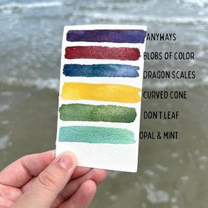 May include: A hand holds a white card with watercolor swatches in shades of purple, red, blue, yellow, green, and teal. Text on the card reads "Anyways, Blobs of Color, Dragon Scales, Curved Cone, Don't Leaf, Opal & Mint."