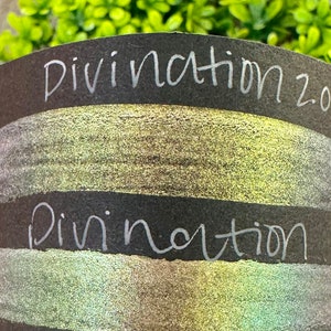 May include: Two swatches of iridescent shimmery paint on a black background. The top swatch is labeled "Divination 2.0" and the bottom swatch is labeled "Divination".