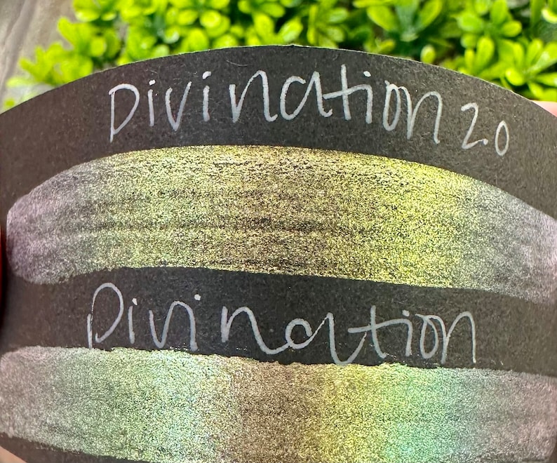 May include: Two swatches of iridescent shimmery paint on a black background. The top swatch is labeled "Divination 2.0" and the bottom swatch is labeled "Divination".