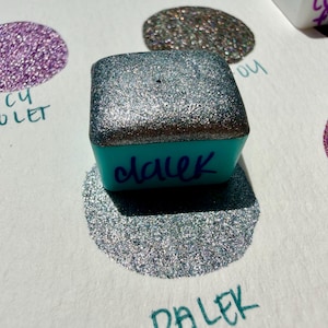 Könnte beinhalten: Ein türkisfarbener Tintenstempel mit dem Wort "dalek" in Schreibschrift. Die Oberseite des Stempels ist mit silbernen Glitzer bedeckt. Unter dem Stempel befindet sich eine silberne Glitzer-Tintenprobe, weitere im Hintergrund.