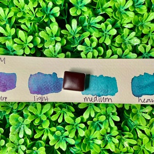 Könnte beinhalten: Eine Aquarellfarbkarte mit vier Farbmustern: Lila, Blau, Türkis und Türkis. Die Karte enthält auch einen kleinen, quadratischen, dunkelbraunen Farbblock. Der Text auf der Karte lautet "NO PRESSURE", "light", "medium" und "heavy".