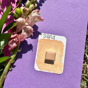 May include: A watercolor paint pan with the word "sand" written on the label. The pan is a light brown color and is sitting on a purple surface.