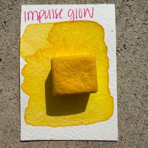 May include: A square, textured, yellow watercolor paint block sits on a white paper with a yellow watercolor wash. The words "impulse glow" are written in pink above the paint block.