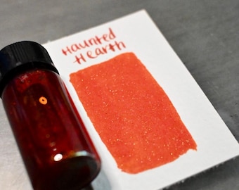 Könnte beinhalten: Eine kleine Glasflasche mit roter Tinte und schwarzem Deckel neben einer weißen Karte mit einem Farbmuster roter Tinte. Auf der Karte steht in roter Tinte "Haunted Hearth".