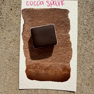 May include: A watercolor swatch on white paper with the words "cocoa spark" in pink script. A dark brown square of watercolor paint sits atop a brown wash, showcasing the color's depth and texture.