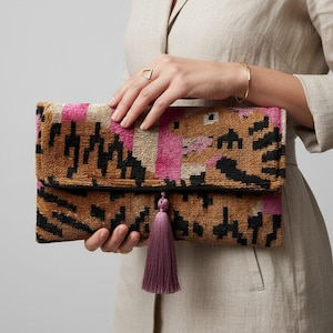 Könnte beinhalten: Eine rechteckige Clutch mit einem strukturierten Webmuster in den Farben Hellbraun, Schwarz und leuchtendem Pink. Die Tasche hat ein Umschlagdesign und ist mit einer langen, herabhängenden mauvefarbenen Quaste an der Vorderseite versehen.