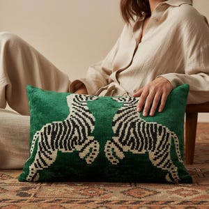 May include: A rectangular throw pillow featuring a deep green velvet fabric with a symmetrical, woven pattern of two cream and black zebras facing each other. The design covers the front surface of the pillow, showcasing a textured, plush finish.