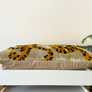 Puede incluir: Una almohada beige con un dise&ntilde;o bordado en negro y dorado de dos tigres. La almohada est&aacute; sobre una estanter&iacute;a blanca.