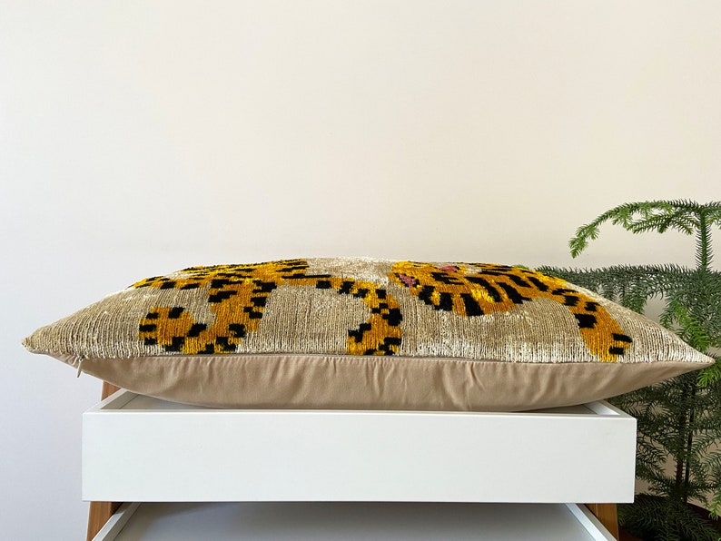Puede incluir: Una almohada beige con un dise&ntilde;o bordado en negro y dorado de dos tigres. La almohada est&aacute; sobre una estanter&iacute;a blanca.