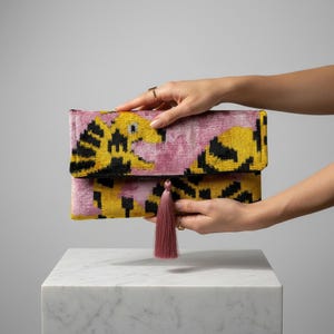 Op de afbeelding: Een roze, gele en zwarte clutch met een bijpassende kwast. De rechthoekige tas heeft een overslagklep. Het ontwerp heeft een herhalend patroon van gele vormen op een roze achtergrond.