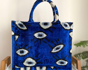 May include: A blue velvet tote bag with a white and blue eye pattern. The bag has two top handles.