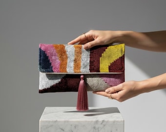Könnte beinhalten: Eine farbenfrohe Clutch mit einem geometrischen Muster in Rosa, Orange, Gelb und Burgunderrot. Die Clutch hat einen Klappverschluss und eine lange, bordeauxfarbene Quaste. Die Tasche wird von einer Person gehalten.