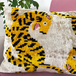 Puede incluir: Una almohada decorativa con fondo blanco y estampado de tigre amarillo y negro. El tigre tiene detalles rosas en la cara.