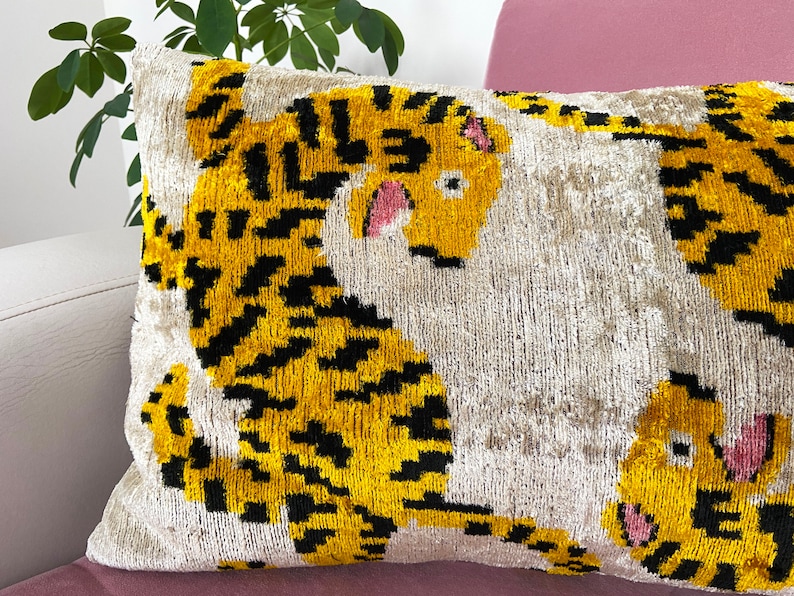 Puede incluir: Una almohada decorativa con fondo blanco y estampado de tigre amarillo y negro. El tigre tiene detalles rosas en la cara.