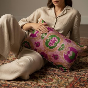 May include: A rectangular throw pillow featuring a vibrant ikat pattern in shades of magenta, bright green, and gold. The fabric displays a repeating ornate motif with stylized floral and pomegranate shapes across the entire surface of the cushion.