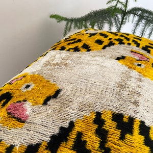 Puede incluir: Una almohada decorativa con fondo blanco y estampado de tigre amarillo y negro. La cara del tigre es prominente con acentos rosados.