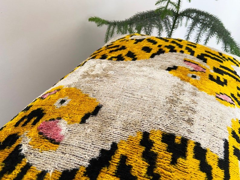 Puede incluir: Una almohada decorativa con fondo blanco y estampado de tigre amarillo y negro. La cara del tigre es prominente con acentos rosados.