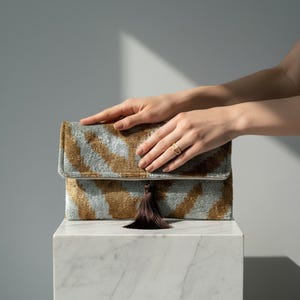Può includere: Pochette rettangolare con chiusura a patta, caratterizzata da un motivo a righe oro e azzurre. La pochette ha un nappa marrone scuro ed è esposta su una superficie di marmo bianco. Le mani di una persona sono delicatamente appoggiate sulla pochette.