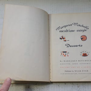 May include: An open vintage cookbook titled Margaret Mitchell's mealtime magic Desserts. The page features black text, red and black illustrations of food, and the author's name, Margaret Mitchell, Director, Home Economics, Volume Two.