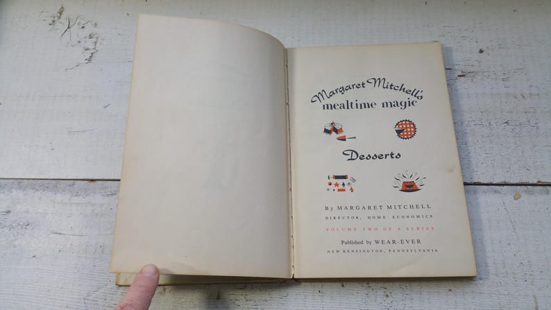 May include: An open vintage cookbook titled Margaret Mitchell's mealtime magic Desserts. The page features black text, red and black illustrations of food, and the author's name, Margaret Mitchell, Director, Home Economics, Volume Two.