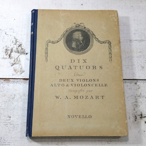 Könnte beinhalten: Ein Vintage-Buchcover mit einem blauen Rücken und einem cremefarbenen Vorderdeckel. Das Cover zeigt ein Porträt eines Mannes in Schwarzweiß, umgeben von einem dekorativen Rahmen. Der Titel lautet "Dix Quatuors Pour Deux Violons Alto & Violoncelle Composés par W.A. Mozart Novello".