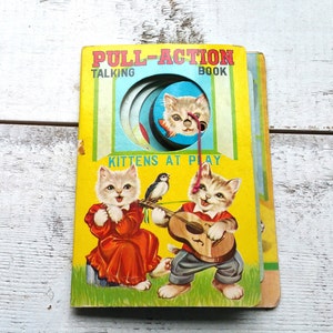 Puede incluir: Un libro parlante de acción de tirar vintage titulado "Pull-Action Talking Book" con las palabras "Kittens at Play" en la portada. El libro presenta una ilustración colorida de dos gatitos tocando música, uno con una guitarra y el otro con un pandero. El libro tiene una lengüeta para tirar que hace un sonido cuando se tira.