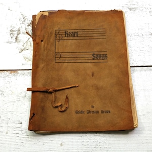 May include: A brown leather-bound book with a title page that reads "Heart Songs" by Goldie Wreston Brown. The book has a leather strap closure.