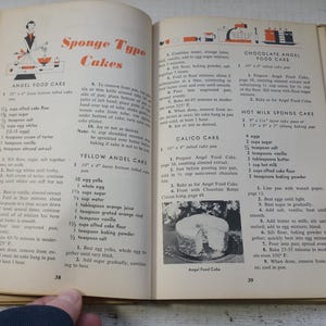 May include: Open vintage cookbook pages with recipes for sponge-type cakes. The pages are yellowed with age and feature illustrations and text. The book is open to pages 38 and 39, with a hand visible at the bottom.